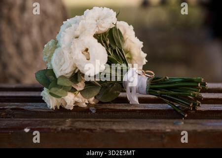 Un gros plan d'un délicat bouquet de fleurs blanches avec des anneaux de mariage en or attachés ensemble sur un banc en bois rustique, capturant romantique et élégant mariage d Banque D'Images