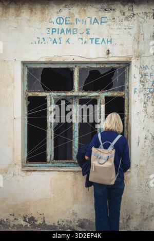 Une femme regarde à travers une fenêtre cassée dans un bâtiment abandonné dans la vieille ville de Marpissa, Paros, Grèce. Banque D'Images