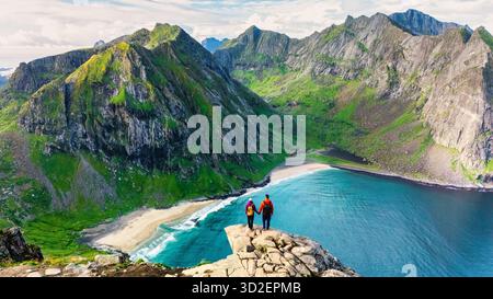 Deux randonneurs admirent le magnifique paysage depuis le pic Ryten, en contemplant les eaux turquoises de la plage de Kvalvika. Ce moment inoubliable capture la beauté du terrain accidenté et de la végétation luxuriante des Lofotens. || autorisation du modèle Banque D'Images