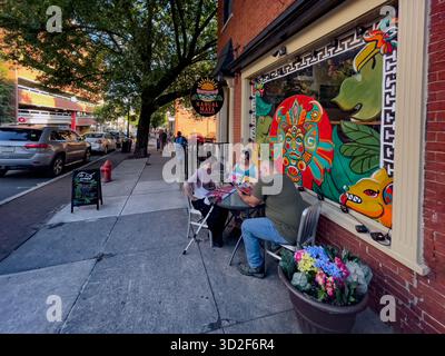 Piétons à un café sur le trottoir sur un coin de rue urbaine verdoyante comme groupe diversifié rassemblé sur le trottoir dans la chaude soirée lig Banque D'Images