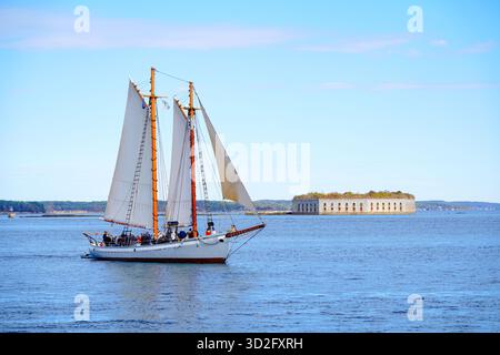 Une goélette à deux mâts passe devant l'historique Fort Gorges dans le port de Portland, dans le Maine, avec des passagers sur le pont profitant des eaux calmes sous un ciel bleu Banque D'Images