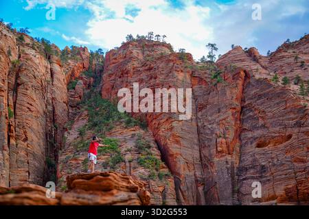 Un randonneur se tient sur un rebord rocheux près d’Angel’s Landing, pointant vers les imposantes falaises de grès qui s’élèvent au-dessus du canyon. Banque D'Images