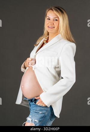 Image studio de la mère attendue en veste blanche et Jean, pose latérale, look maternité paisible et confiant Banque D'Images