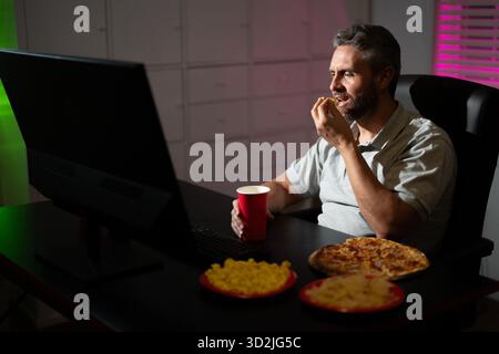 Homme d'affaires manger de la pizza en heures supplémentaires. Homme occupé mangeant le dîner et travaillant en ligne au bureau de nuit. Entrepreneur millénaire travaille tard dans la nuit tout en profitant Banque D'Images