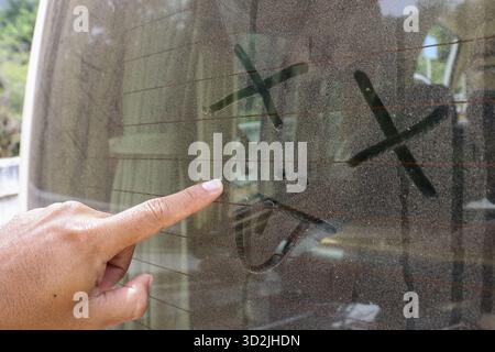 La main ludique pointe le doigt sur le dessin espiègle du visage triste sur la fenêtre poussiéreuse de voiture. Ce message gribouillé sur une surface de verre sale montre une idée créative Banque D'Images