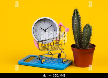 Temps de magasinage. Chariot de supermarché avec horloge, calculatrice et cactus sur fond jaune Banque D'Images