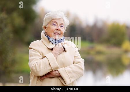 Belle femme âgée souriante avec les cheveux gris portant un manteau beige élégant posant à l'extérieur dans le parc d'automne. Femme senior à la mode appréciant la nature Banque D'Images