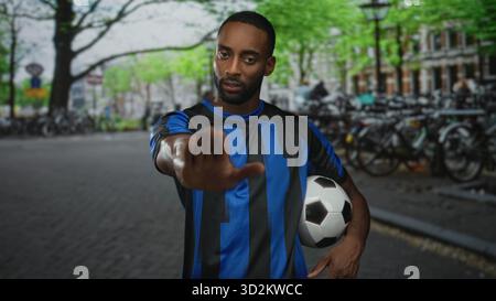 Jeune homme afro-américain en maillot bleu tient le ballon de football et montre un geste d'arrêt de paume dans la rue de la ville ; détermination de soutien de l'équipe. Banque D'Images