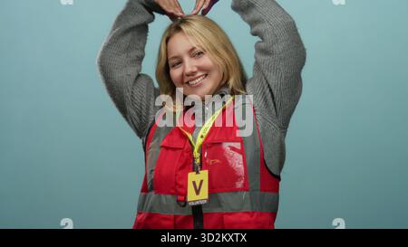 Femme blonde souriante en gilet de volontaire rouge fait symbole de coeur avec des mains sur fond de mur bleu. Banque D'Images