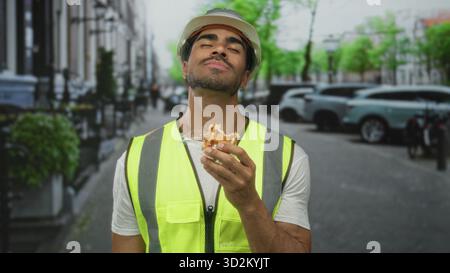 Homme mangeant de la gaufre dorée dans une rue urbaine animée portant un gilet jaune et un casque d'ingénieur ; satisfaction. Banque D'Images
