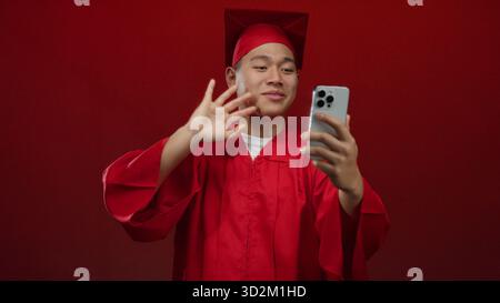 Jeune homme chinois étudiant en robe de graduation rouge faisant un appel vidéo sur smartphone au-dessus d'un mur rouge isolé célébrant la réussite scolaire avec une joyf Banque D'Images