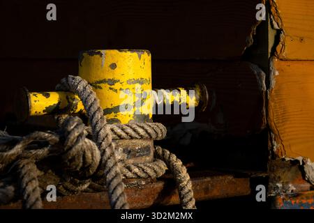 Borne jaune vieillie avec corde épaisse fixant le bateau en bois vieilli, avec peinture écaillée et métal rouillé sur une structure à quai Banque D'Images