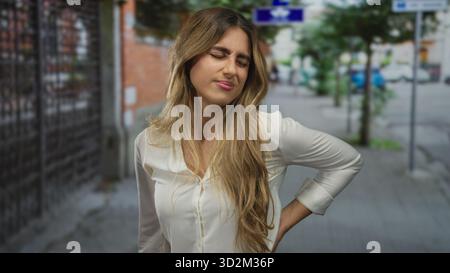 Jeune femme blonde en chemisier blanc tient le bas du dos sur la rue pavée ; mal de dos inconfort douleur de stress. Banque D'Images