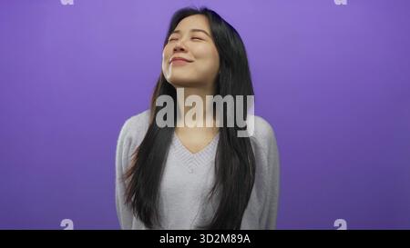 Femme souriant avec les yeux fermés sur un fond violet vif, mettant en valeur l'expression sereine et le charme jeune. Banque D'Images