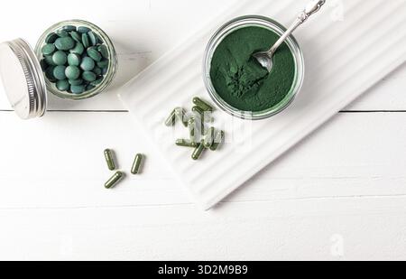 Spiruline en poudre, comprimés, capsules dans divers pots sur fond en bois blanc et support en céramique. Vue de dessus. Suppléments nutritionnels sains Banque D'Images