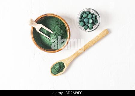 Bol en bois, cuillère et cuillère remplis de poudre d'algues spiruline verte sur fond blanc. Un pot en verre avec des comprimés de spiruline. Vue de dessus. Détox Banque D'Images