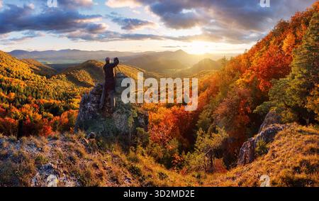 Homme sur la pierre sur la colline et belle vallée de montagne dans la brume au coucher du soleil coloré en automne. Guy, montagne forêt rouge, ciel dramatique avec soleil à l'automne. Banque D'Images