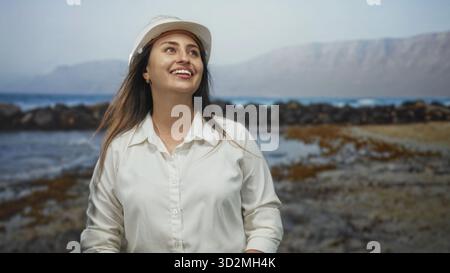 Femme portant un casque de protection sourit et regarde à travers le rivage rocheux sur la plage ; optimisme professionnel côtier. Banque D'Images