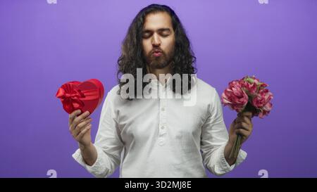 Homme souriant tout en tenant une boîte cadeau coeur rouge et un bouquet rose dans un studio violet ; geste romantique. Banque D'Images