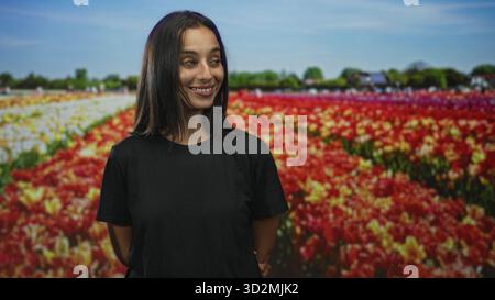 Femme souriant avec les yeux fermés debout au milieu de rangées de tulipes colorées dans un champ avec les mains derrière son dos et chemise noire décontractée ; sérénité. Banque D'Images