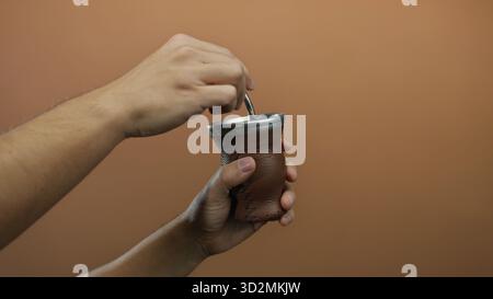 Homme préparant la boisson traditionnelle de maté en utilisant une gourde en cuir brun sur un fond de mur isolé. Banque D'Images