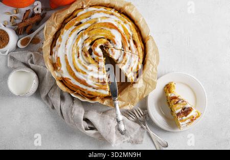 Vue de dessus d'un rouleau géant de citrouille à la cannelle avec glaçage à la crème au beurre et un morceau coupé sur l'assiette à proximité, table blanche. Photo de haute qualité, espace de copie. Banque D'Images