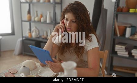 Femme tient le téléphone à l'oreille tout en lisant une tablette et en disposant des vases en céramique sur la table dans un studio ; concentration. Banque D'Images