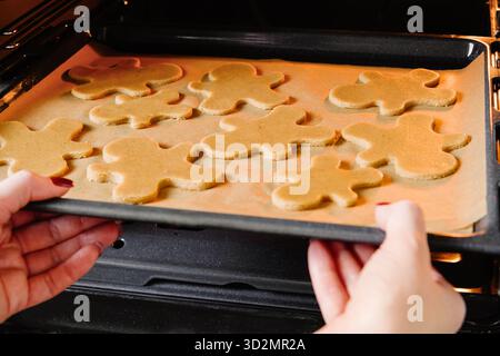Les mains féminines mettent un plateau de cuisson avec l'homme de biscuit en pain d'épices de Noël dans le four dans la cuisine. Fond de concept de cuisson de Noël. Banque D'Images