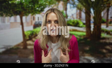 Femme tient le doigt tout en pointant vers elle-même dans le parc où la jeune blonde se tient au milieu du vert à l'extérieur avec une expression surprise. Banque D'Images