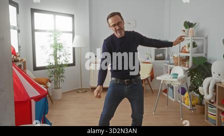 Homme dansant joyeusement dans une crèche colorée remplie de jouets et de meubles, mettant en valeur une atmosphère intérieure heureuse et animée. Banque D'Images