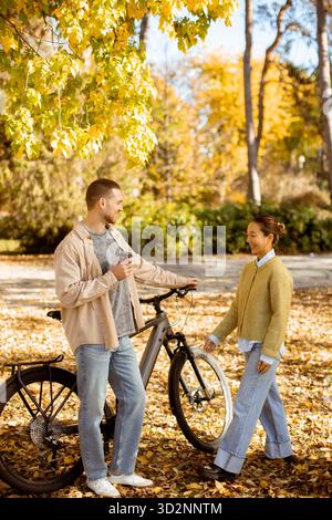 Un jeune couple multiracial partage un moment joyeux dans un parc animé, entouré de feuilles d'automne colorées tout en profitant d'une journée ensoleillée ensemble. Banque D'Images