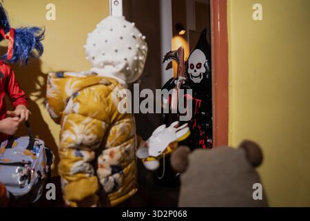 Les enfants habillés de costumes créatifs d'Halloween frappant à la porte pour rencontrer la personne dans la tenue de faucheuse sinistre tenant faux pendant le tour du soir festif ou t Banque D'Images