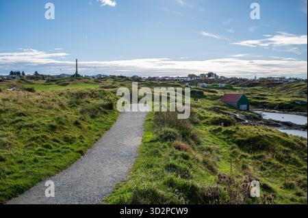 Kyststien, Haugesund, Norvège avec Haraldshaugen, chemin de gravier serpente à travers des collines verdoyantes, menant à une petite maison verte par un plan d'eau sous un cl Banque D'Images