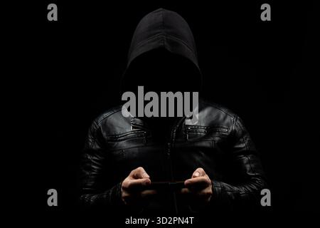 Gros plan sur un homme hacker à capuche sans visage tenant un téléphone portable en forme horizontale avec les deux mains et un fond noir Banque D'Images