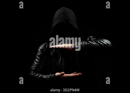 Gros plan sur un homme à capuche sans visage montrant un espace spécifique avec ses deux mains devant lui et un fond noir Banque D'Images