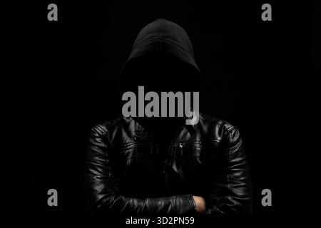 Gros plan sur un homme hacker à capuche sans visage dans l'ombre avec les bras croisés et un fond noir Banque D'Images