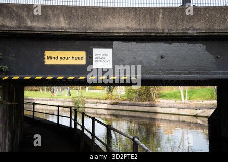 Deux panneaux de sécurité sur un pont bas au-dessus de la rivière Lea navigation près de Waltham Cross – un jaune « Mind Your Head » et un blanc « cyclistes s’il vous plaît descendre ». Banque D'Images