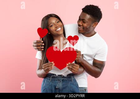 Jeune couple noir partage l'affection tout en tenant Red Paper Hearts sur un fond rose Banque D'Images