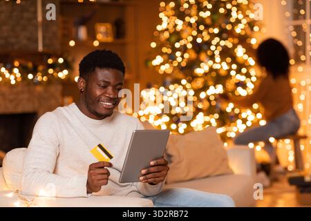 Millennial Black Man aime les achats en ligne tandis que sa femme décore l'arbre de Noël pour le nouvel an Banque D'Images