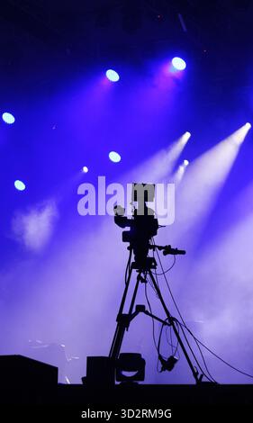 Silhouette d'une caméra vidéo professionnelle sur un trépied à côté d'une scène avec un éclairage bleu et violet dramatique Banque D'Images
