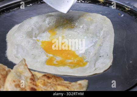 Délicieux gros plan du chef préparant du pain plat roti salé sur une poêle chaude. Cook ajoute des œufs frais à la pâte pour un petit déjeuner de rue asiatique savoureux et populaire Banque D'Images