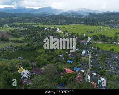 La vue enchanteresse du paysage aérien capture un village rural paisible niché dans une vallée verdoyante, entouré de montagnes majestueuses. le toit doré du temple ajoute Banque D'Images