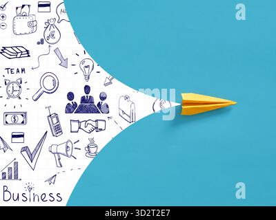 Avion de papier jaune volant, portant des idées d'affaires et des icônes dessinées sur le bloc-notes, sur fond bleu. Démarrage d'entreprise, progrès et concept d'innovation Banque D'Images