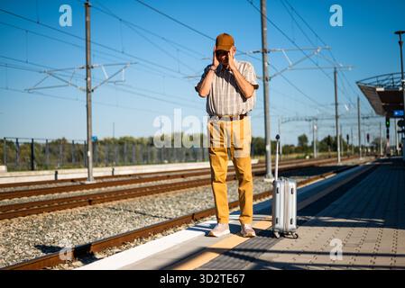 Un homme senior se tient debout à la gare. Il a raté le départ du train et a tenu la tête dans l'incrédulité. Personnes âgées voyageant. Banque D'Images
