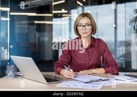 Femme d'affaires asiatique confiante dans un bureau d'entreprise moderne examinant des documents à son bureau avec un ordinateur portable et des lunettes, prêt et concentré sur la planification financière et la signature de contrat Banque D'Images