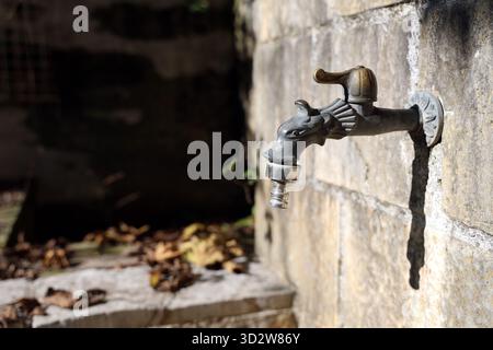 Un vieux robinet extérieur en métal dépasse d'un mur de pierre altérée, projetant une longue ombre en plein soleil. Idéal pour jardin, cour et maint extérieur Banque D'Images