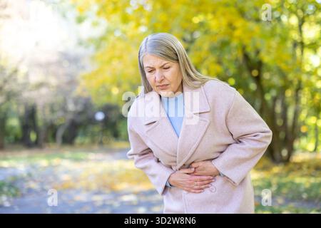 Femme âgée souffrant de douleurs abdominales sévères, tenant son ventre avec les deux mains tout en se tenant debout dans un parc avec des arbres d'automne en arrière-plan, exprimant l'inconfort Banque D'Images
