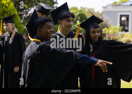 Diplômant les étudiants en casquettes et robes, célébrant à l'extérieur et pointant avec enthousiasme. Obtention du diplôme, éducation, réalisation, célébration, commencement, réussite || autorisation du modèle Banque D'Images