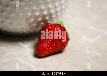 Plan détaillé d'une fraise rouge mûre reposant près d'un bol en céramique blanche, mettant l'accent sur la fraîcheur naturelle. Banque D'Images