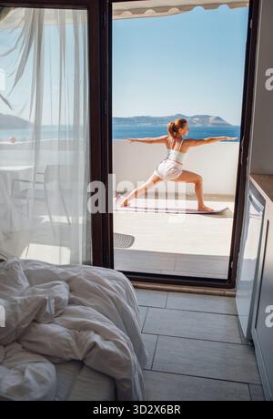 Femme d'âge moyen faisant le yoga du matin guerrier exercice d'équilibre de posture sur la vue mer Terace chambre d'hôtel, faisant Virabhadrasana pose qui rend votre corps mor Banque D'Images